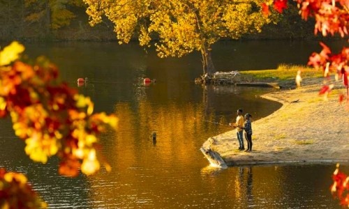 Two people fishing by a serene lake surrounded by vibrant autumn foliage.