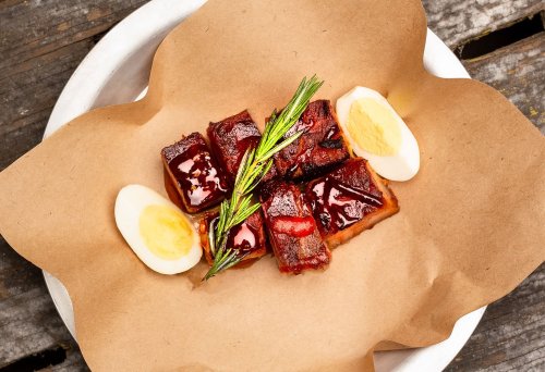 A plate with glazed meat cubes, garnished with rosemary and served with halved boiled eggs on brown paper.