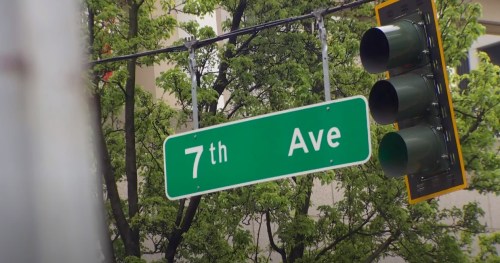 Green street sign for "7th Ave" with traffic lights and trees in the background.