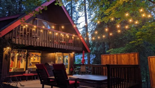 A cozy cabin in the woods, illuminated by string lights, with a deck and hot tub surrounded by trees.