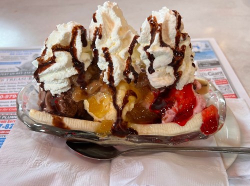 A colorful banana split topped with whipped cream, chocolate syrup, and various fruit toppings in a glass dish.
