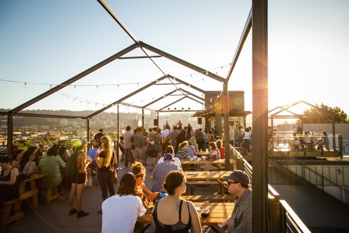 A lively rooftop gathering at sunset, with people socializing and enjoying the view from a wooden deck.