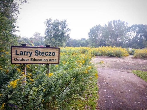 Sign for "Larry Steczo Outdoor Education Area" with a path and wildflowers in a natural setting.