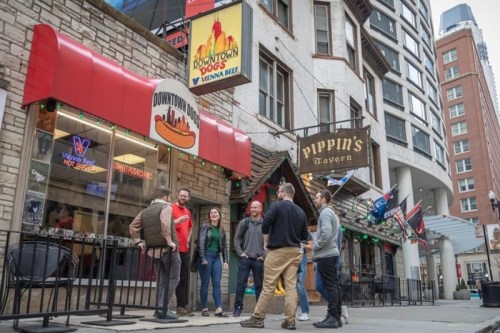 A group of people chatting outside Pippin's Tavern and Downtown Dogs in an urban setting.
