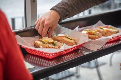 A hand reaches for a hot dog topped with condiments, placed in a red basket alongside other hot dogs.