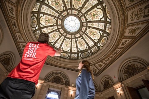 Two people admire a beautifully designed stained glass dome in a historic building. One points at the ceiling.