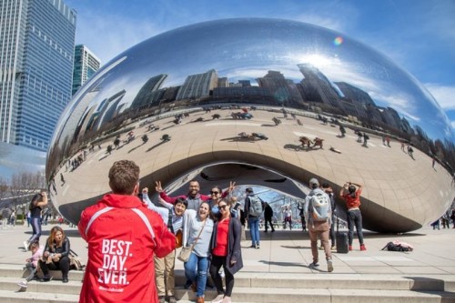 A group of people poses in front of a large, reflective sculpture in a city park, with buildings and visitors in the background.
