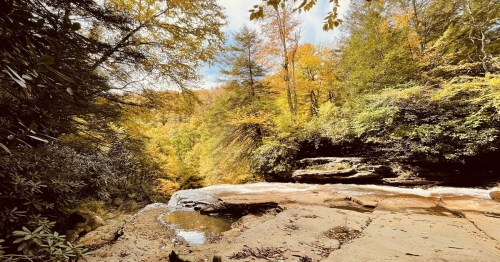 A serene landscape featuring a rocky riverbank surrounded by vibrant autumn foliage and a clear blue sky.