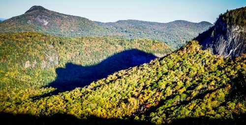 A scenic view of rolling hills covered in autumn foliage, with long shadows cast by the mountains in the background.