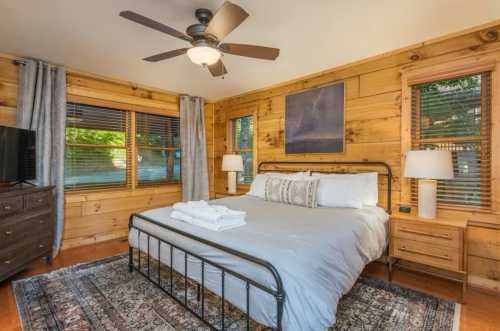 Cozy bedroom with wooden walls, a queen bed, two lamps, a dresser, and large windows letting in natural light.