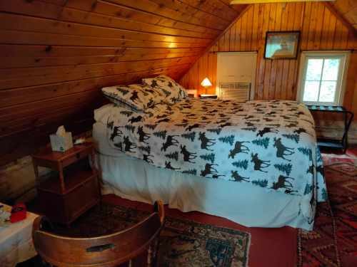 Cozy attic bedroom with wooden walls, a bed with moose-patterned bedding, a small table, and a window.