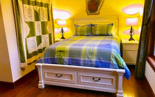 Cozy bedroom featuring a white bed with a plaid blanket, two lamps, and a quilted curtain. Warm yellow walls.