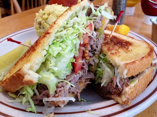 A close-up of a sandwich filled with lettuce, tomato, and meat, served with a side of pickles and a scoop of potato salad.