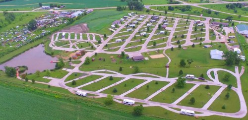Aerial view of a campground with RVs, roads, and a pond, surrounded by green fields and trees.