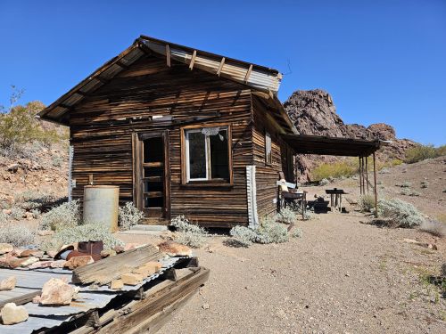 A rustic wooden cabin in a desert landscape, surrounded by rocks and sparse vegetation under a clear blue sky.