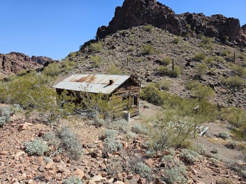 Abandoned wooden cabin with a rusty roof, surrounded by rocky terrain and sparse desert vegetation under a clear blue sky.