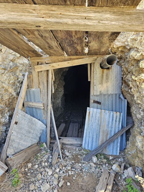 An abandoned mine entrance with wooden beams and metal sheets, surrounded by rocky terrain.