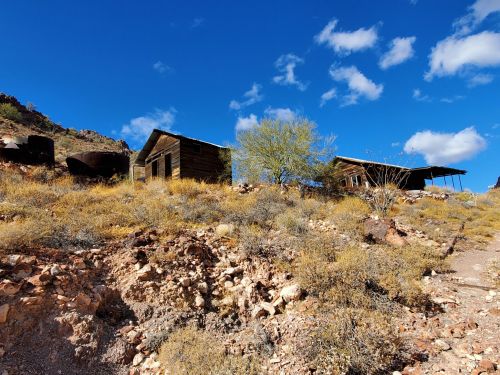 Abandoned wooden buildings on a hillside, surrounded by dry grass and shrubs under a blue sky with scattered clouds.