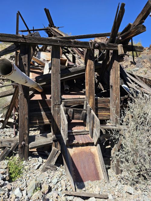 A dilapidated wooden structure with broken beams and debris, set against a clear blue sky and rocky terrain.