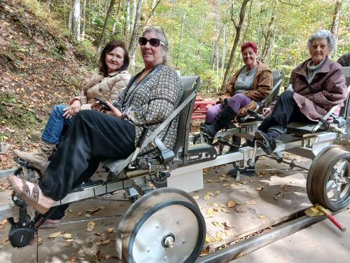 Four women sitting on a unique bike-like vehicle in a forested area, enjoying a sunny day outdoors.