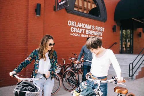 Two women with bicycles stand outside a craft brewery, smiling and looking at their bikes. A man is in the background.
