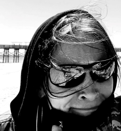 A person with long hair and sunglasses stands on a beach, with a pier visible in the background and wind blowing.