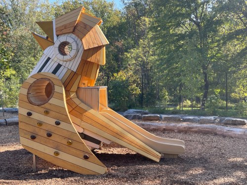 A wooden playground structure shaped like a bird, featuring slides and climbing elements, surrounded by trees.