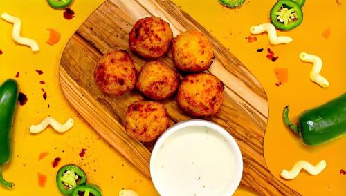 A wooden board with six golden-brown balls and a small bowl of white sauce, surrounded by green peppers and colorful toppings.