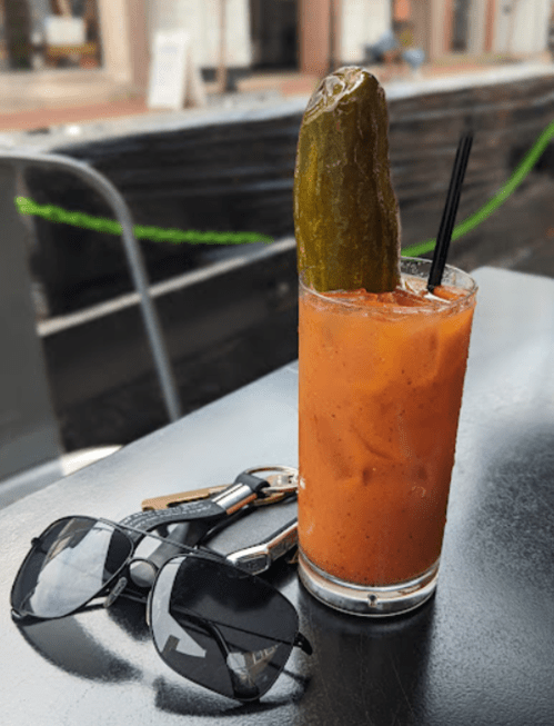 A glass of Bloody Mary with a pickle garnish, sunglasses, and keys on a table.