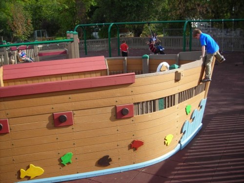 A playground ship structure with children playing, one boy climbing on the side, colorful fish shapes on the hull.