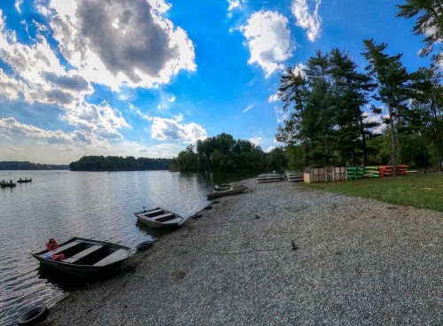 A serene lakeside scene with boats lined up on a gravel shore, surrounded by trees and a blue sky with clouds.