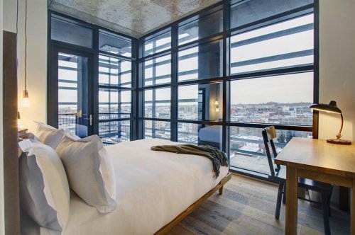 Modern bedroom with large windows, a cozy bed, and a wooden desk, offering a view of the city skyline.