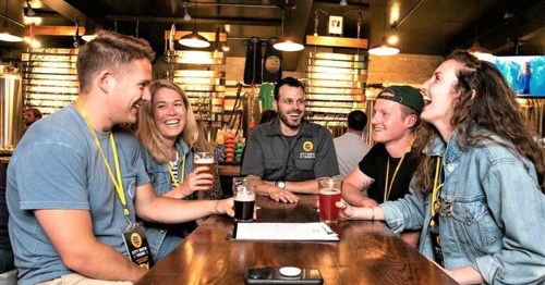 A group of five friends enjoying drinks and laughter at a brewery, with a bartender in the background.