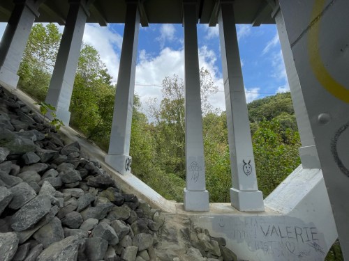 View from under a bridge, showcasing tall concrete pillars and a rocky area with greenery and blue sky in the background.