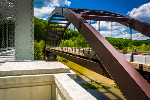 A modern arch bridge spans a river, surrounded by lush greenery and a bright blue sky with fluffy clouds.