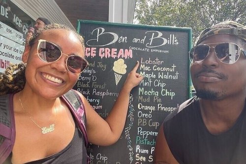 Two smiling people pose in front of a chalkboard menu for Ben and Bill's Ice Cream, showcasing various flavors.