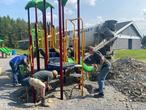 Workers installing a playground structure, with a concrete mixer in the background and construction materials nearby.