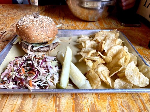 A sesame seed burger on a tray with coleslaw, pickles, and potato chips on a wooden table.