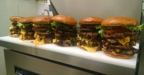 A row of towering burgers stacked with multiple patties, cheese, lettuce, and tomato on toasted buns.