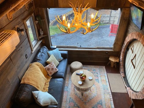 Cozy living space with a leather sofa, rustic decor, a chandelier, and a round table with fruit, surrounded by large windows.