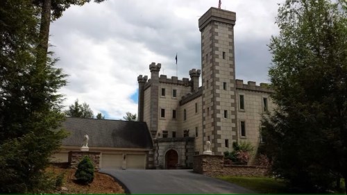 A stone castle with a tall tower, surrounded by trees and a landscaped yard, under a cloudy sky.