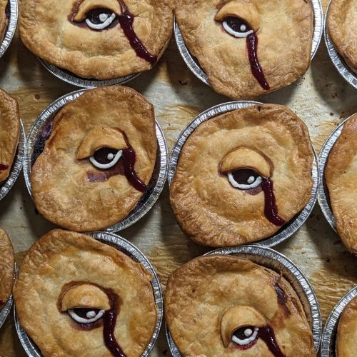 A collection of pie crusts with artistic designs featuring eyes and red filling, arranged in circular tins.