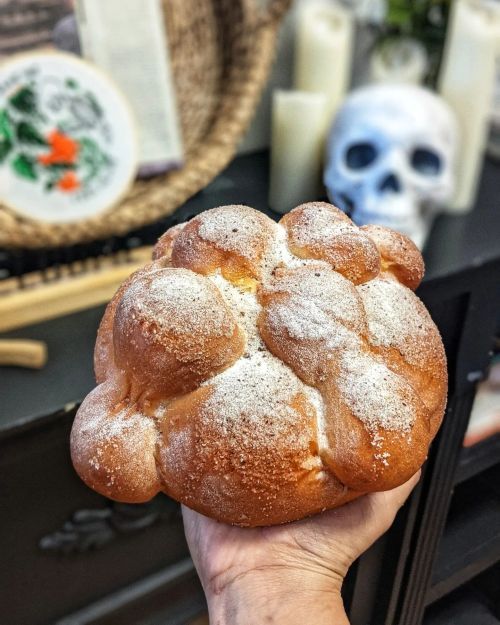 A hand holding a round, fluffy bread topped with powdered sugar, with candles and a skull in the background.