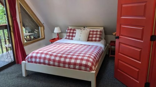 Cozy bedroom with a checkered red and white bedspread, a lamp, and a bright red door. Large windows with a view.