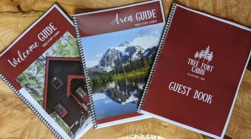Three spiral-bound booklets: "Welcome Guide," "Area Guide," and "Guest Book," featuring a cabin and mountain scenery.