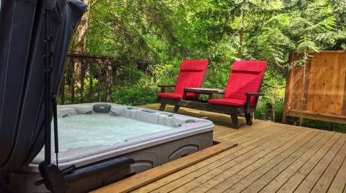 A hot tub on a wooden deck surrounded by greenery, with two red chairs and a small table nearby.