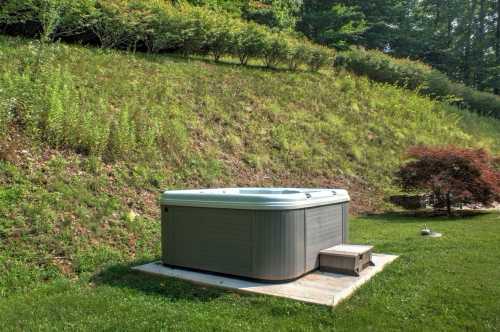 A hot tub sits on a concrete pad in a grassy area, surrounded by lush greenery and a sloped hillside.