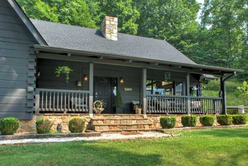 A cozy gray house with a stone chimney, front porch, and landscaped yard surrounded by trees.