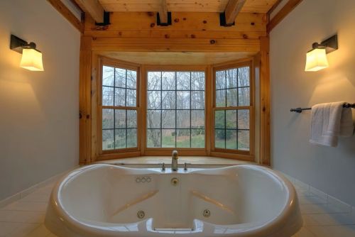 A spacious bathroom featuring a large jacuzzi tub under a wooden beam ceiling, with windows overlooking a natural view.