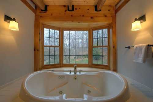 A spacious bathroom featuring a large jacuzzi tub under a wooden beam ceiling, with windows overlooking a natural view.